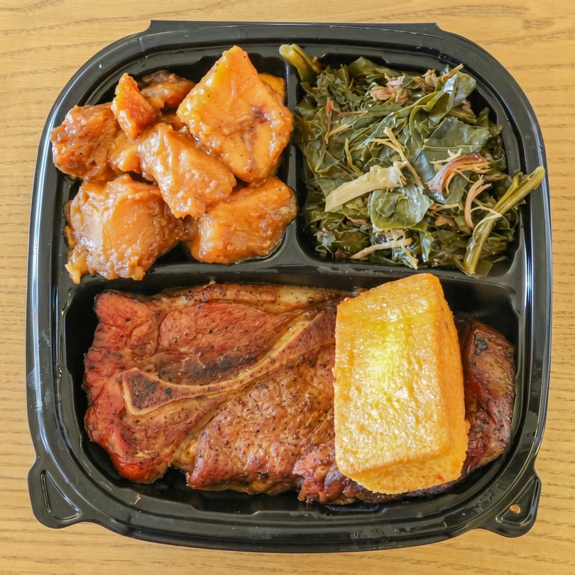 A takeout container with a pork chop, cornbread, candied yams, and collard greens.