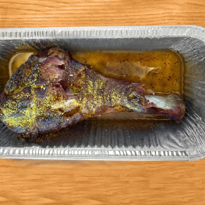 Roasted lamb shank with yellow seasoning in a foil pan with broth, on a wooden table.