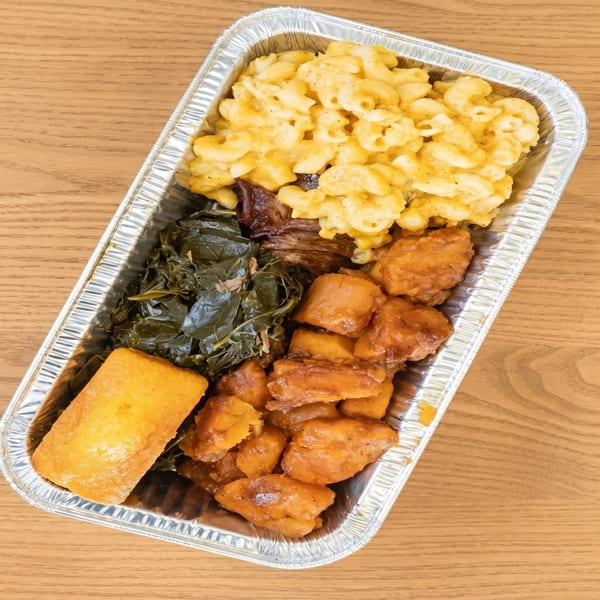 A takeout container filled with macaroni and cheese, collard greens, sweet potatoes, and cornbread.