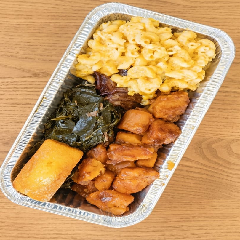 A takeout container filled with macaroni and cheese, collard greens, sweet potatoes, and cornbread.
