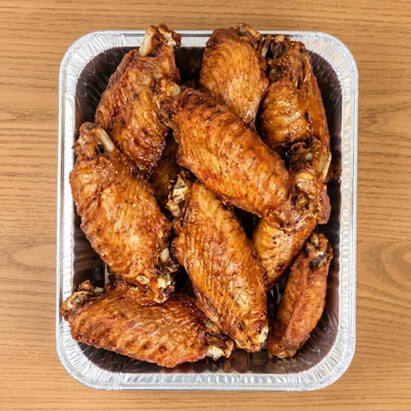 A foil tray filled with numerous glazed, dark brown chicken wings on a wooden table.