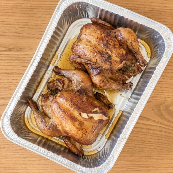 Two whole roasted chickens with crispy skin and seasoning in a foil pan.